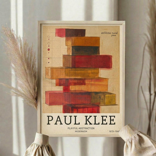 May include: Framed abstract art print with overlapping geometric rectangles in red, orange, brown, and yellow tones on cream background, displayed on wooden surface next to dried pampas grass stems and neutral fabric with gathered details in soft natural lighting.