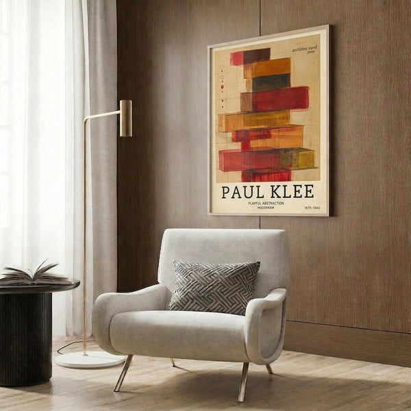 May include: Paul Klee abstract art poster with colorful geometric rectangles in red, orange, brown and yellow tones, displayed in light wood frame on wood-paneled wall. Modern reading nook features cream upholstered armchair with geometric patterned pillow, brass floor lamp with white marble base, and dark round side table with open book.
