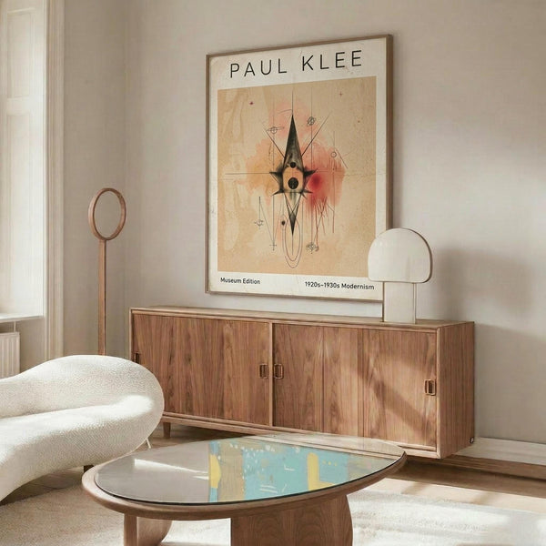 May include: Paul Klee modernist art print in light wood frame hanging above walnut sideboard in bright living room. Abstract artwork features geometric shapes, lines and warm earth tones on cream background. White curved chair and oval glass coffee table with colorful surface visible in foreground. Modern floor lamp with circular shade stands beside sideboard.