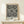 May include: Framed modern art poster featuring black polka dot pattern on white background with "YAYOI KUSAMA JAPAN 19" text at bottom, displayed on light wood console table next to small wrapped gift with evergreen sprig, against cream colored wall