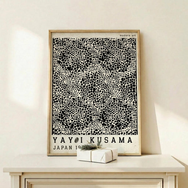 May include: Framed modern art poster featuring black polka dot pattern on white background with "YAYOI KUSAMA JAPAN 19" text at bottom, displayed on light wood console table next to small wrapped gift with evergreen sprig, against cream colored wall