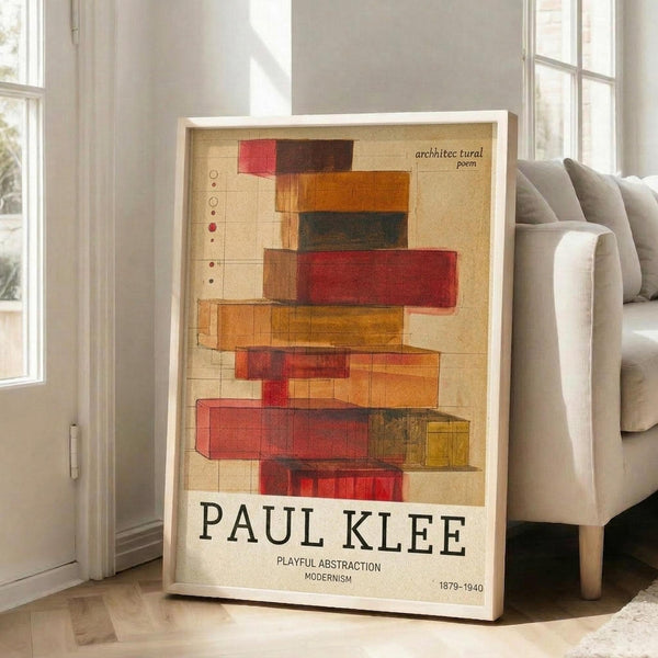 May include: Framed poster featuring abstract geometric composition with overlapping translucent rectangles in warm reds, oranges, yellows and browns on cream background. Text reads "architectural poem" at top and "PAUL KLEE PLAYFUL ABSTRACTION MODERNISM 1879-1940" at bottom. White frame leans against white wall next to beige sofa in bright sunlit room with large windows and hardwood floors.