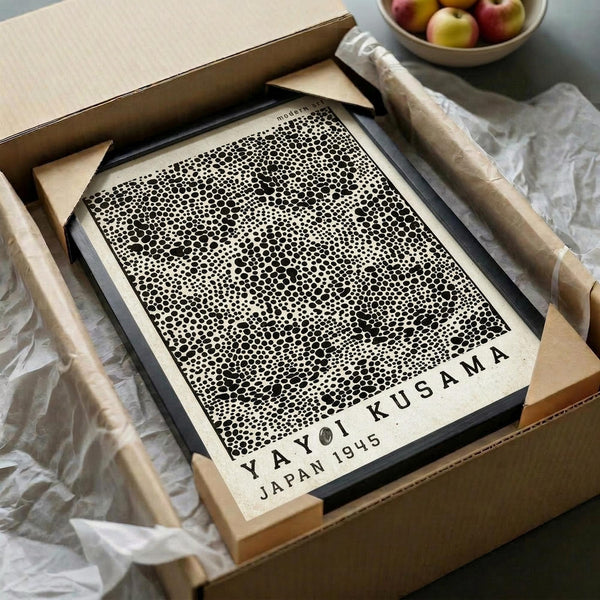May include: Framed black and white dotted pattern art print in wooden frame, displayed in minimalist setting with kraft paper packaging, wooden dowels, white tissue paper, and bowl of apples in background. The artwork features dense polka dot design with "YAYOI KUSAMA JAPAN 1945" text at bottom.