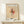 May include: Framed Paul Klee abstract modernist artwork print displayed on light wood surface against white wall. The poster features geometric shapes in black, pink, and beige with linear elements on aged paper background, housed in natural wood frame. Small white wrapped gift with evergreen sprig sits beside the frame.