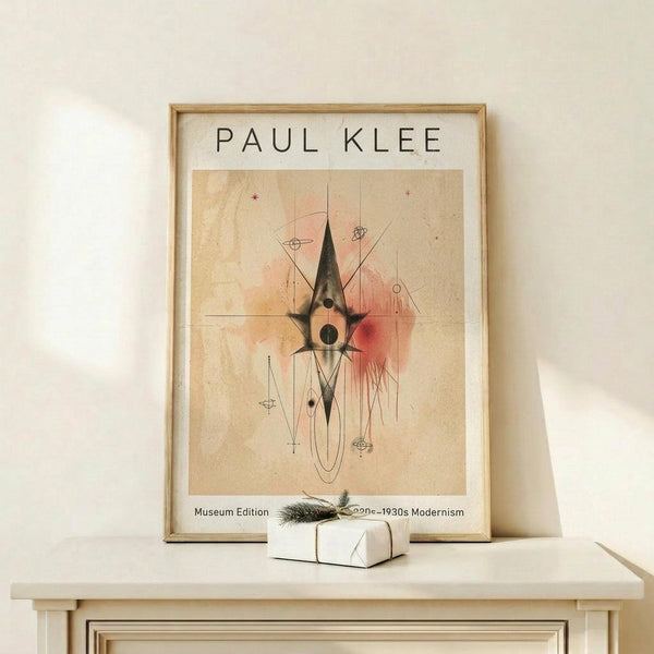 May include: Framed Paul Klee abstract modernist artwork print displayed on light wood surface against white wall. The poster features geometric shapes in black, pink, and beige with linear elements on aged paper background, housed in natural wood frame. Small white wrapped gift with evergreen sprig sits beside the frame.