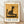 May include: Framed poster print featuring Paul Klee's 1924 modernist artwork of a stylized black cat sitting on a chair against golden yellow background. The minimalist geometric composition shows bold black shapes forming the cat and furniture. Print has white matting in light wood frame, displayed on wooden floor against white wall with soft shadows.