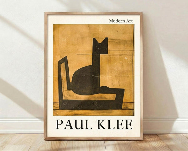 May include: Framed poster print featuring Paul Klee's 1924 modernist artwork of a stylized black cat sitting on a chair against golden yellow background. The minimalist geometric composition shows bold black shapes forming the cat and furniture. Print has white matting in light wood frame, displayed on wooden floor against white wall with soft shadows.