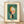 May include: Framed abstract art print leaning against white wall on light wood floor. Cream matted poster features geometric composition with orange crescent, white and coral semicircles, black rectangular forms on teal-green background. Text identifies artist, artwork title, and 1924 date. Natural lighting creates soft shadows on textured white wall.