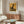 May include: A framed modern art poster featuring a stylized black cat silhouette on golden yellow background, displayed above a mid-century wooden sideboard in a minimalist living room. The room includes a white curved lounge chair, round glass coffee table, and copper floor lamp, creating a warm, contemporary interior setting with neutral wall colors and natural lighting.