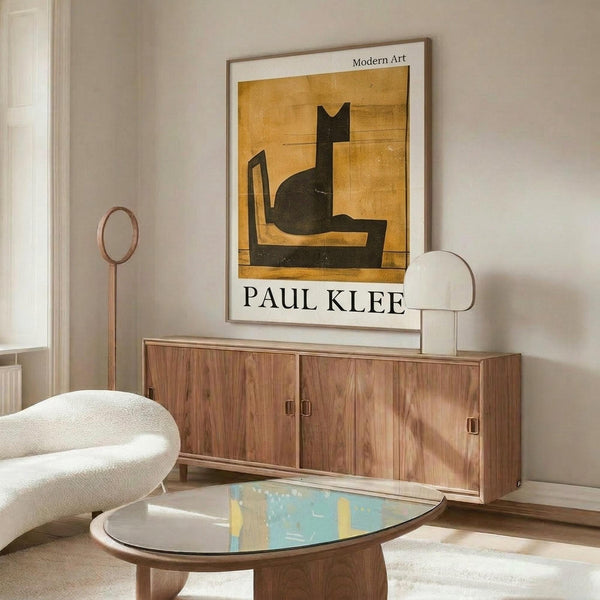 May include: A framed modern art poster featuring a stylized black cat silhouette on golden yellow background, displayed above a mid-century wooden sideboard in a minimalist living room. The room includes a white curved lounge chair, round glass coffee table, and copper floor lamp, creating a warm, contemporary interior setting with neutral wall colors and natural lighting.