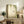 May include: Framed botanical artwork featuring green hanging ferns and a small hummingbird on cream background, displayed on rustic wooden sideboard alongside potted fern plant, ceramic vases, and books in neutral-toned living room with beige armchair and soft natural lighting