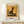 May include: Framed art print displaying a minimalist black cat silhouette sitting on a chair against warm golden-yellow background, styled as vintage exhibition poster with "Modern Art" and "PAUL KLEE" text below. Light wood frame rests on pale wooden surface beside small wrapped gift with evergreen sprig against cream wall.