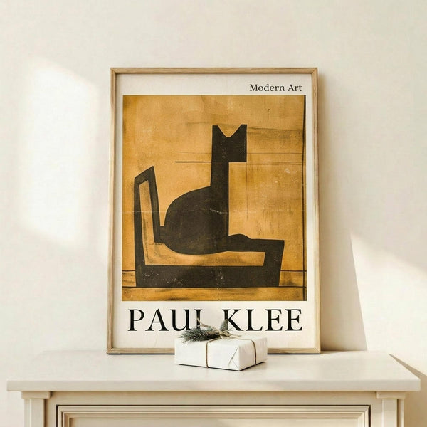 May include: Framed art print displaying a minimalist black cat silhouette sitting on a chair against warm golden-yellow background, styled as vintage exhibition poster with "Modern Art" and "PAUL KLEE" text below. Light wood frame rests on pale wooden surface beside small wrapped gift with evergreen sprig against cream wall.