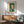 May include: Framed Kandinsky abstract art print hanging above wooden sideboard in modern living room. Poster features geometric shapes in green, orange, white and pink tones. Mid-century furniture includes teal upholstered armchair with wooden frame, walnut credenza with sliding doors, white ceramic table lamp, and round coffee table. Natural sunlight creates shadows on white walls.