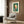May include: Framed Kandinsky abstract art print hanging on wood-paneled wall above modern beige armchair with geometric pillow. Artwork features colorful circular and angular shapes in green, orange, white, and pink. Room styled with brass floor lamp, round side table with open book, and warm neutral tones throughout.