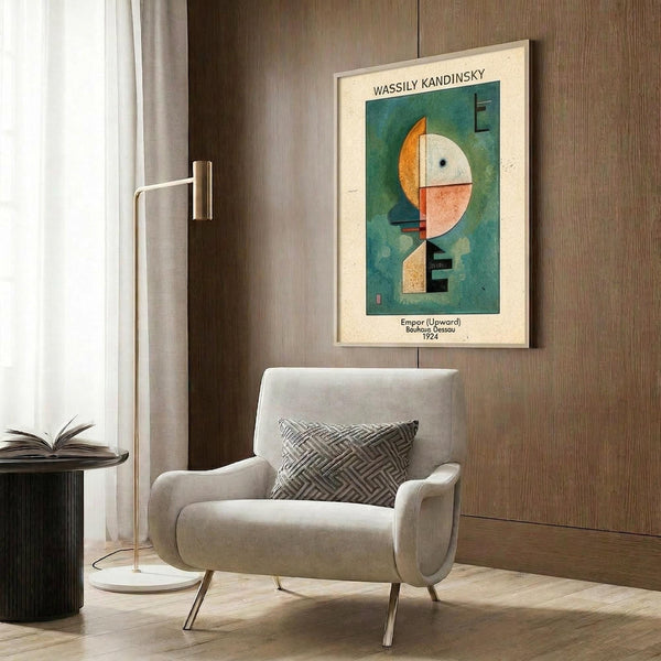 May include: Framed Kandinsky abstract art print hanging on wood-paneled wall above modern beige armchair with geometric pillow. Artwork features colorful circular and angular shapes in green, orange, white, and pink. Room styled with brass floor lamp, round side table with open book, and warm neutral tones throughout.