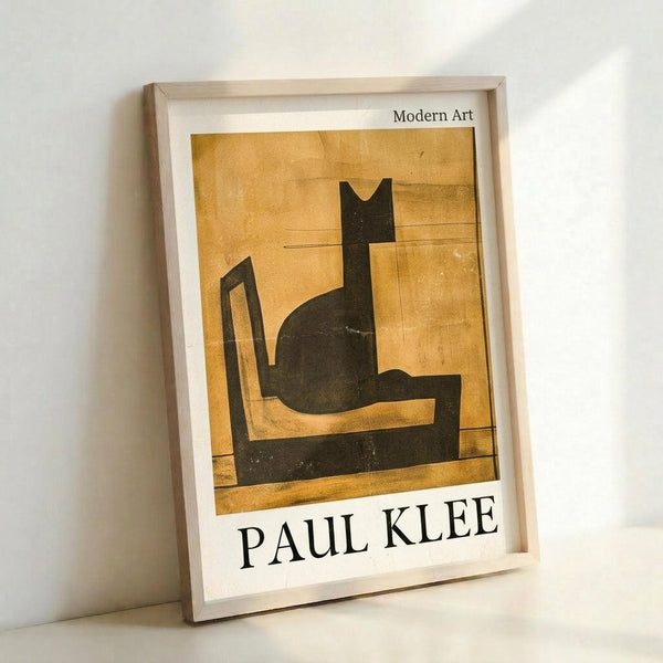 May include: Framed poster print leaning against white wall showing Paul Klee's modernist artwork of a stylized black cat silhouette sitting on geometric shapes against warm golden yellow background. Light wood frame with white matting displays the vintage-style art print with "PAUL KLEE" text at bottom and "Modern Art" at top.