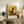 May include: Framed poster with golden yellow background featuring black silhouette of a sitting cat, displayed on rustic wooden sideboard alongside fern plant, ceramic vases, and books in warm-toned living room setting with cream armchair and neutral walls.
