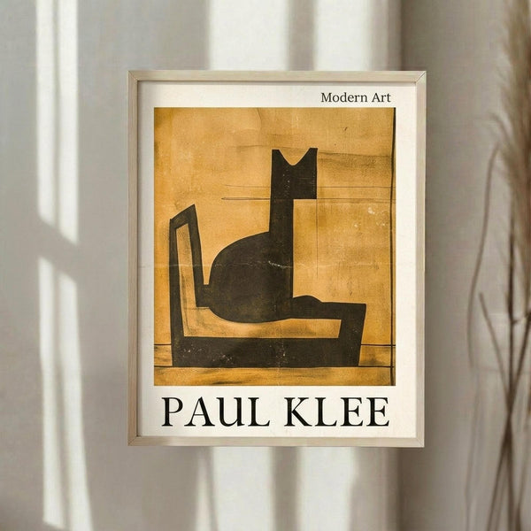 May include: Framed art print featuring Paul Klee's abstract black cat silhouette on warm golden-yellow background, displayed on white wall with natural lighting creating soft shadows. Print shows geometric, modernist style with bold black shapes forming stylized feline figure. Light wooden frame with white matting complements neutral interior setting.