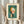 May include: Person in cream clothing holding framed abstract art print featuring geometric shapes in orange, white, and coral against teal-green background. Dried wheat stems visible in left hand. Print shows Kandinsky's modernist composition with circles, rectangles, and linear elements. Neutral indoor setting with soft natural lighting.