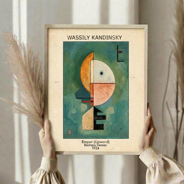 May include: Person in cream clothing holding framed abstract art print featuring geometric shapes in orange, white, and coral against teal-green background. Dried wheat stems visible in left hand. Print shows Kandinsky's modernist composition with circles, rectangles, and linear elements. Neutral indoor setting with soft natural lighting.