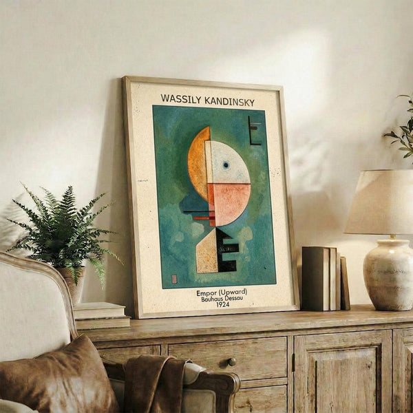 May include: Framed Kandinsky abstract art print featuring geometric shapes in teal, orange, white and pink on cream mat, displayed on rustic wooden sideboard with fern plant, ceramic vase, books, and brown leather chair visible in cozy living room setting