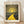 May include: Framed art print leaning against white wall on light wood floor. Print features dotted pattern artwork with bright yellow winding path leading to glowing sun, surrounded by dark landscape with colorful circular elements. Text reads "YAYOI KUSAMA" at top and "Modern Art 1944" at bottom. Natural wood frame, cream colored mat border, soft natural lighting creates gentle shadows on textured white wall.