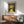 May include: Framed poster print featuring Yayoi Kusama artwork with yellow winding path leading to bright sun against dark dotted sky, displayed above mid-century modern wooden sideboard with white ceramic lamp, next to sage green upholstered armchair in sunlit room with white walls.