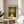 May include: Framed art print displayed on wooden round side table next to beige upholstered chair. Print features yellow winding path leading to bright sun with radiating circles against dark dotted sky. Artwork has cream background with "YAYOI KUSAMA" at top and "Modern Art 1944" at bottom. Natural wood frame, soft window lighting, and partial plant leaves visible in corner of minimalist room setting.