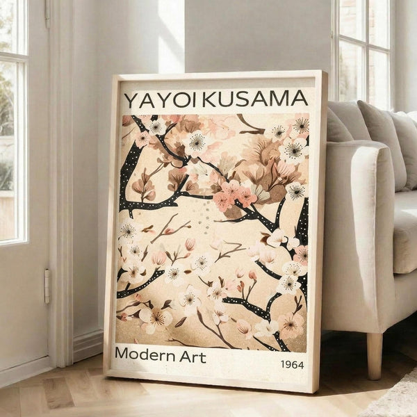 May include: Large framed art print leaning against white wall next to beige upholstered chair in bright living room. Print features cherry blossom branches with pink and white flowers on cream background, displaying "YAYOI KUSAMA" at top and "Modern Art 1964" at bottom. Natural wood frame, sunlight streaming through windows onto hardwood floors.