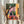May include: Person holding framed colorful poster print featuring figure in plaid shirt walking through yellow archway toward pink garden area with large tree and foliage. Artwork has vibrant orange, pink, green, and yellow colors with "Take it Slow" text overlay. Thin light wood frame, hands wearing cream ruffled sleeves visible, dried wheat stems in background against white curtains.