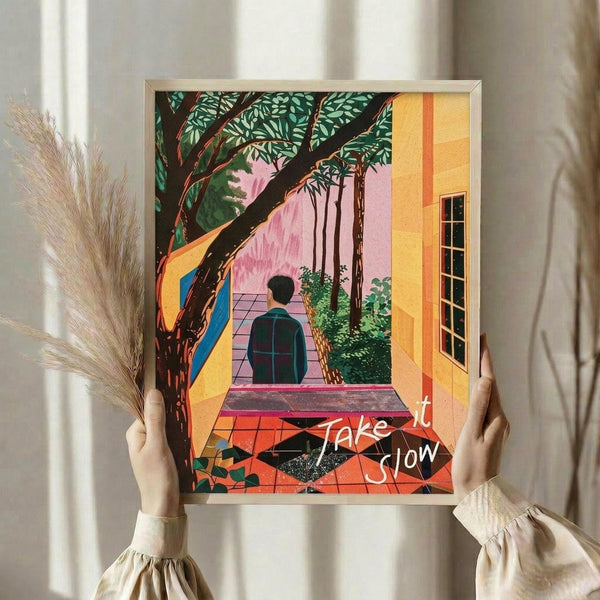May include: Person holding framed colorful poster print featuring figure in plaid shirt walking through yellow archway toward pink garden area with large tree and foliage. Artwork has vibrant orange, pink, green, and yellow colors with "Take it Slow" text overlay. Thin light wood frame, hands wearing cream ruffled sleeves visible, dried wheat stems in background against white curtains.
