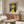 May include: Framed art print hanging above wooden sideboard in modern living room. Poster shows yellow winding path leading to bright sun against dark dotted sky. White curved chair and glass coffee table visible. Mid-century modern furniture with warm wood tones. Natural lighting creates shadows on cream walls.