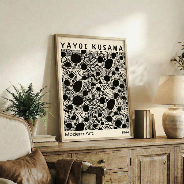 May include: Framed black and white polka dot art print leaning against white wall on rustic wooden sideboard. Print features dense pattern of various sized black circles and dots on cream background. Scene includes beige upholstered chair, potted fern, stacked books, ceramic vase, and warm natural lighting creating cozy living room atmosphere.