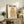 May include: Vintage Japanese poster featuring geometric orange owl design with Japanese text, displayed in light wood frame on rustic wooden console. Room styled with neutral tones, fern plant in woven basket, small olive tree in ceramic pot, leather throw pillow on beige chair. Warm natural lighting creates cozy living space atmosphere.