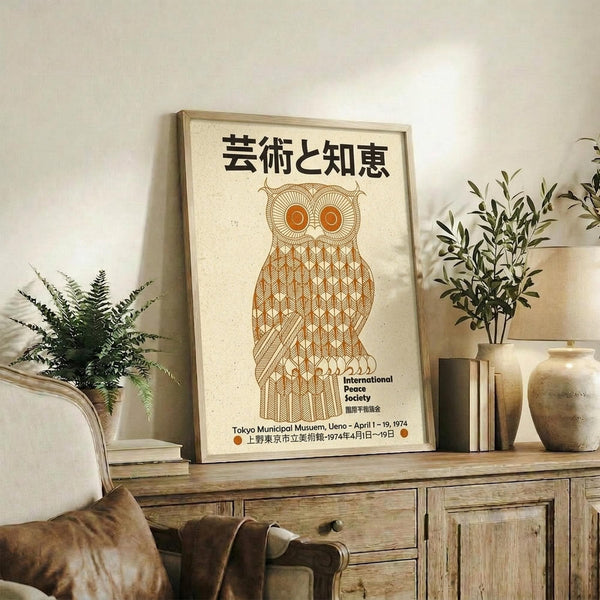 May include: Vintage Japanese poster featuring geometric orange owl design with Japanese text, displayed in light wood frame on rustic wooden console. Room styled with neutral tones, fern plant in woven basket, small olive tree in ceramic pot, leather throw pillow on beige chair. Warm natural lighting creates cozy living space atmosphere.