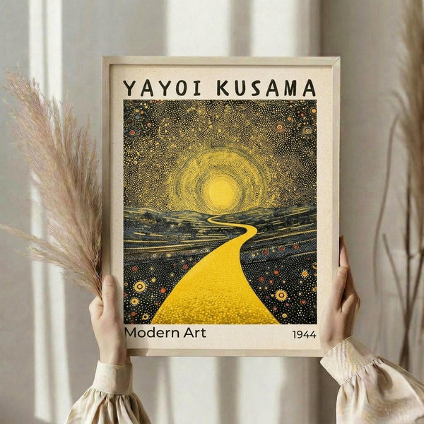 May include: Person holding framed art print featuring dotted yellow sun radiating concentric circles over landscape with winding golden path through dark starry field. Pointillism style artwork in light wood frame against white curtained window. Dried pampas grass stems visible on left side.