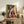 May include: Framed poster print displayed on rustic wooden sideboard in living room setting. Artwork shows figure sitting on tiled steps under large tree with green foliage, surrounded by warm yellow, pink and orange architectural elements. White cursive text reads "Take it Slow" at bottom. Scene includes fern plant, ceramic vase, books, and beige armchair with leather cushion.
