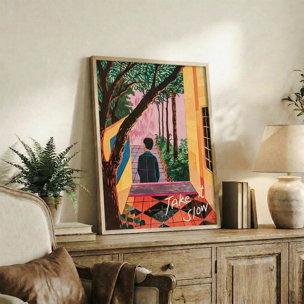 May include: Framed poster print displayed on rustic wooden sideboard in living room setting. Artwork shows figure sitting on tiled steps under large tree with green foliage, surrounded by warm yellow, pink and orange architectural elements. White cursive text reads "Take it Slow" at bottom. Scene includes fern plant, ceramic vase, books, and beige armchair with leather cushion.