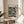 May include: Framed poster with black polka dot pattern on cream background displayed on round wooden side table next to beige upholstered chair. Natural lighting creates soft shadows on neutral wall. Green plant leaves visible in corner. Print features dense arrangement of various sized black circles and dots with text at top and bottom.