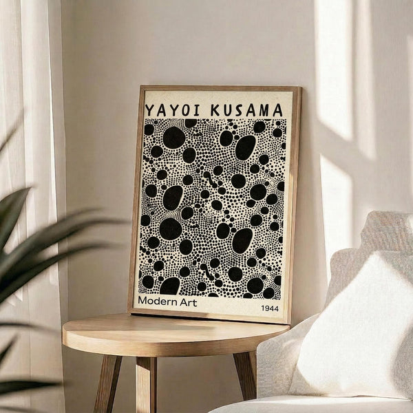 May include: Framed poster with black polka dot pattern on cream background displayed on round wooden side table next to beige upholstered chair. Natural lighting creates soft shadows on neutral wall. Green plant leaves visible in corner. Print features dense arrangement of various sized black circles and dots with text at top and bottom.