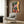 May include: Colorful poster print hanging on wood-paneled wall above beige upholstered armchair with geometric pillow. Artwork shows figure walking up steps through tropical garden scene with large tree, bright yellow and pink buildings, decorative tilework, and red script text overlay. Modern reading nook setting with brass floor lamp and side table.