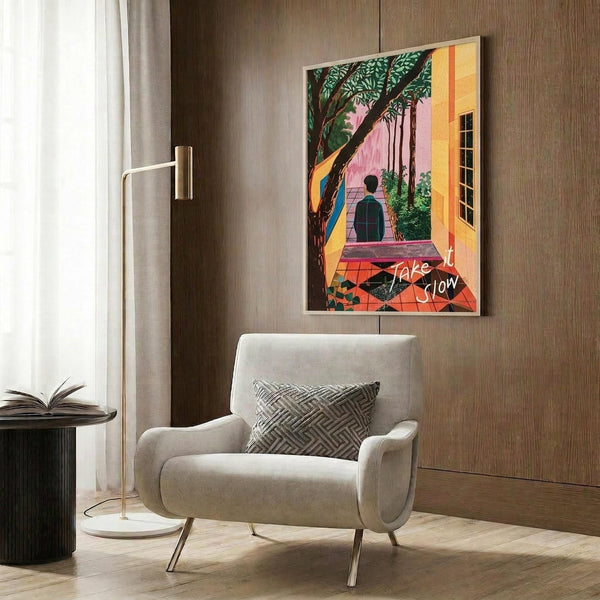 May include: Colorful poster print hanging on wood-paneled wall above beige upholstered armchair with geometric pillow. Artwork shows figure walking up steps through tropical garden scene with large tree, bright yellow and pink buildings, decorative tilework, and red script text overlay. Modern reading nook setting with brass floor lamp and side table.