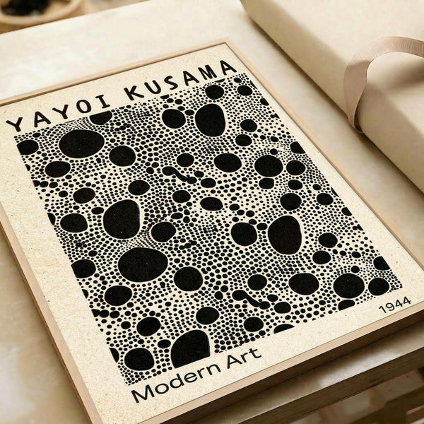 A black and white abstract art print featuring various sized polka dots scattered across a cream-colored background. The artwork displays Yayoi Kusama's signature dot pattern with circles ranging from tiny to large, creating a dense, organic composition. Text reads "YAYOI KUSAMA" at the top and "Modern Art" with "1944" at the bottom. The print appears on textured paper within a light wooden frame, photographed on a neutral surface with soft lighting.