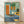 May include: Framed abstract geometric art print featuring a vintage turntable on a blue textured surface surrounded by colorful rectangular shapes in orange, green, and teal. The composition includes circular elements in pink, blue, and cream, with French text at the bottom reading "Douce nuit sous les étoiles". The artwork has a mid-century modern aesthetic with layered geometric forms and muted vintage colors, displayed in a light wood frame against a white wall on wooden flooring.