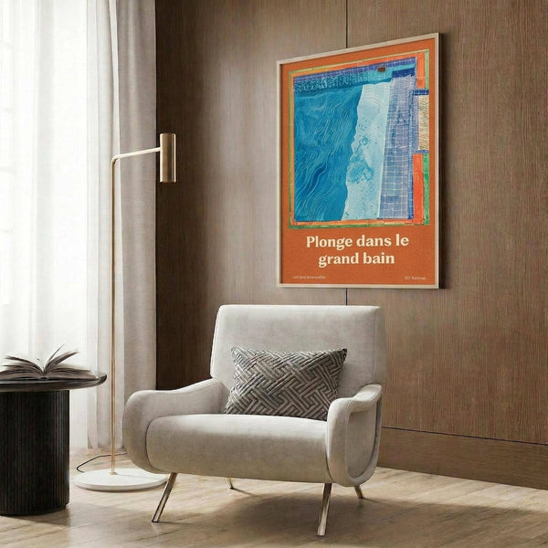 May include: Modern living room scene with beige armchair and geometric pillow beneath wood paneled wall. Orange-framed poster displays abstract blue swimming pool artwork with French text "Plonge dans le grand bain." Gold floor lamp and dark round side table with open book complete the contemporary interior setting.