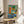 May include: Colorful abstract art print featuring geometric shapes in blue, orange, and green tones with a vinyl record player centerpiece. The framed poster sits on a round wooden side table next to a beige upholstered chair in a bright, minimalist room with white walls and natural lighting. Plant leaves visible in corner add organic contrast to the modern geometric artwork.