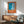 May include: Framed poster with blue and orange abstract design featuring pool-like imagery and French text "Plonge dans le grand bain" displayed above mid-century modern wood credenza with ceramic table lamp. Green upholstered armchair with wooden frame sits nearby in sunlit room with white walls and wooden floors.