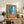 May include: Framed poster with orange background and blue swimming pool illustration, featuring French text "Plonge dans le grand bain." Displayed on rustic wooden sideboard alongside fern plant, ceramic vase, lamp, and books in neutral-toned living room setting with beige armchair.