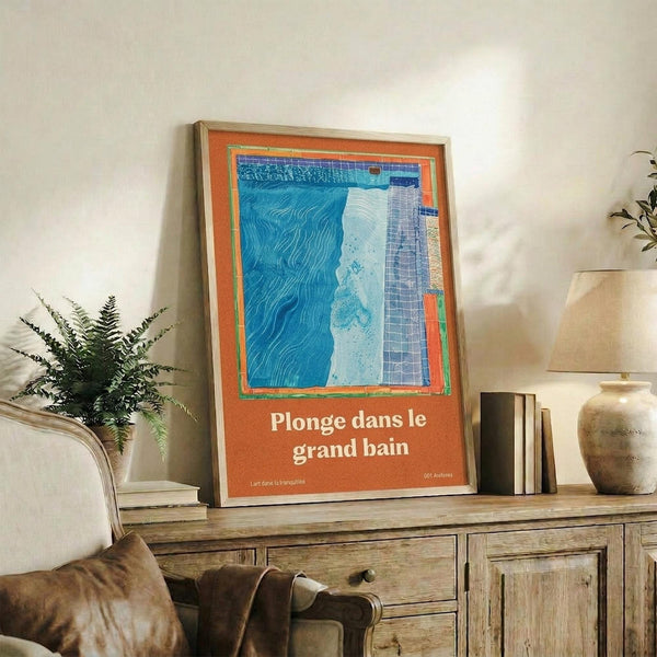 May include: Framed poster with orange background and blue swimming pool illustration, featuring French text "Plonge dans le grand bain." Displayed on rustic wooden sideboard alongside fern plant, ceramic vase, lamp, and books in neutral-toned living room setting with beige armchair.